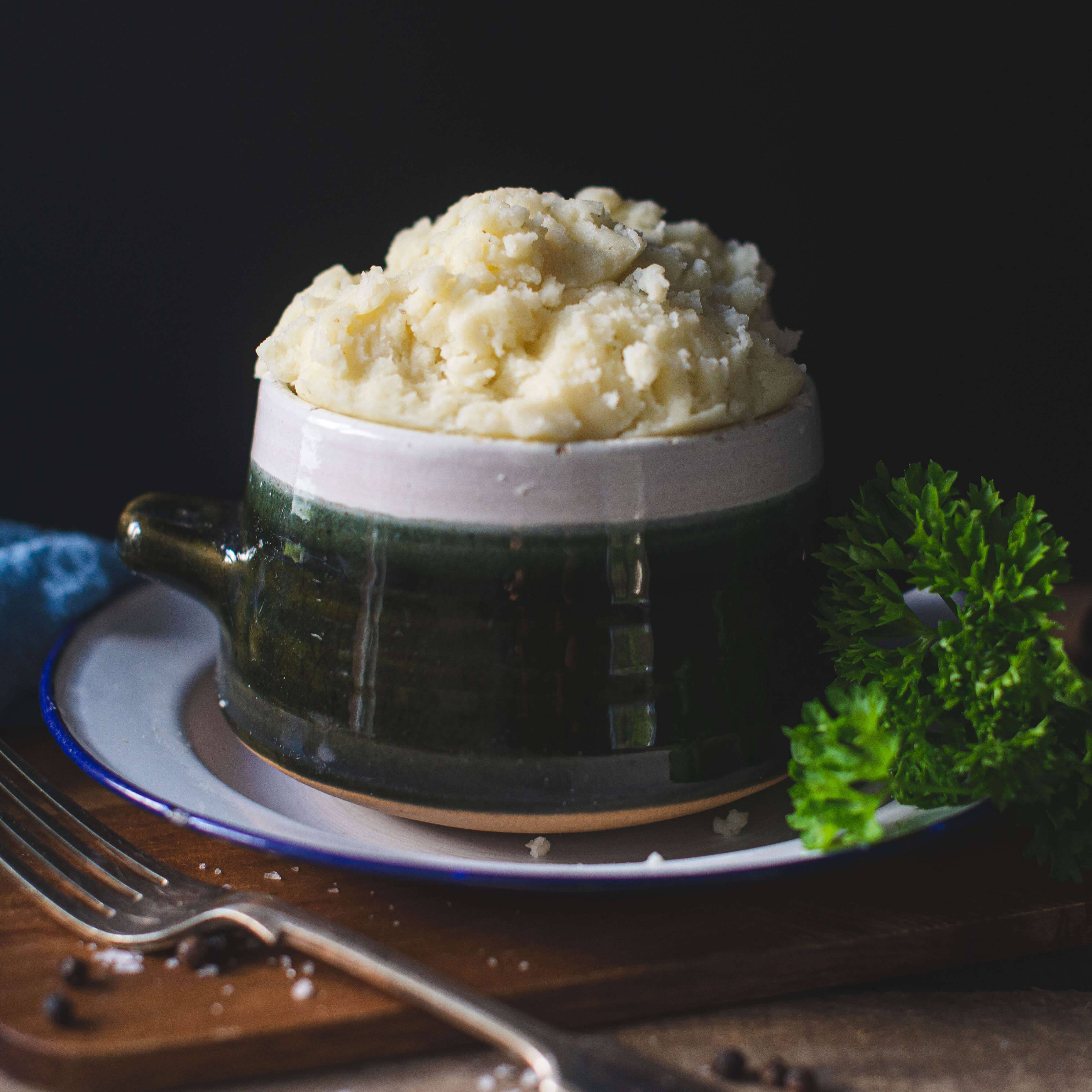 Mud Pies’ Buttery Mashed Potato in a green and white bowl, topped with parsley and peppercorns, sits on a white plate with a fork.