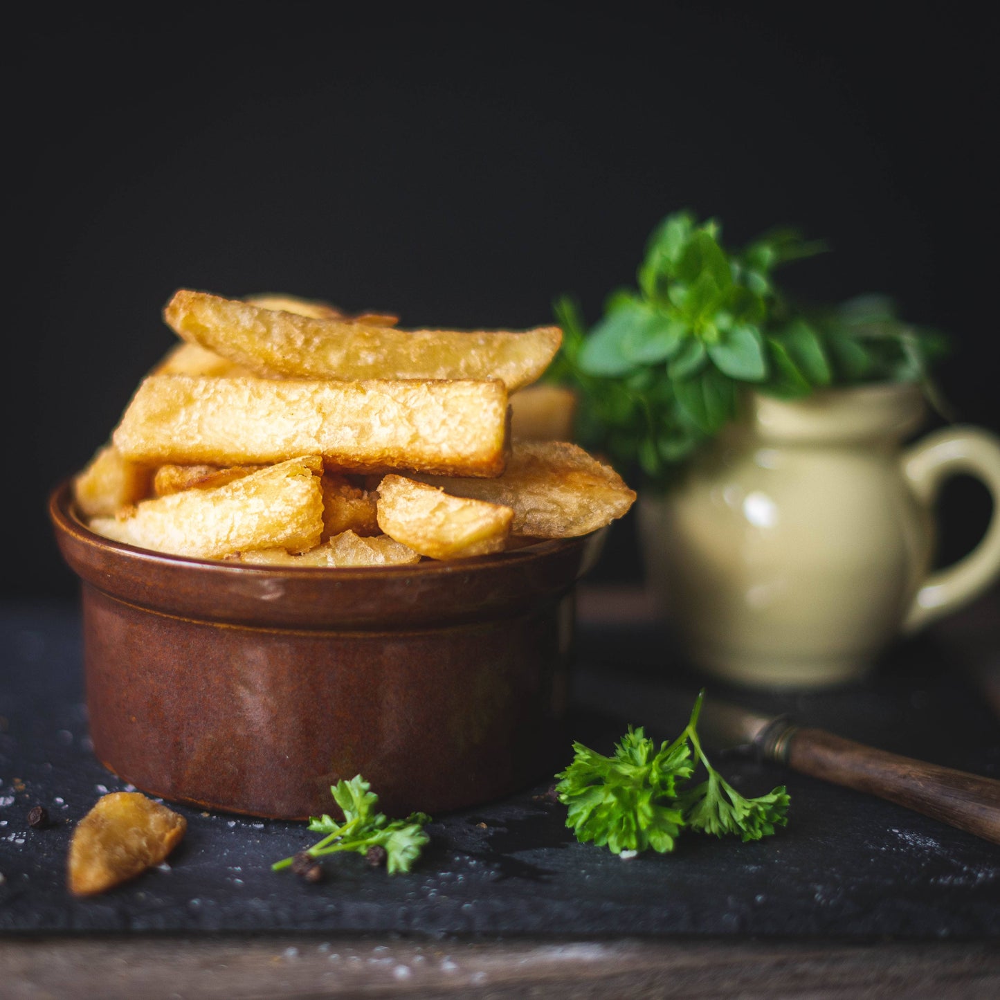 Mud Pies Beer Battered Chips in a brown ceramic bowl rest on a dark surface, with parsley and a cream jug of herbs behind.