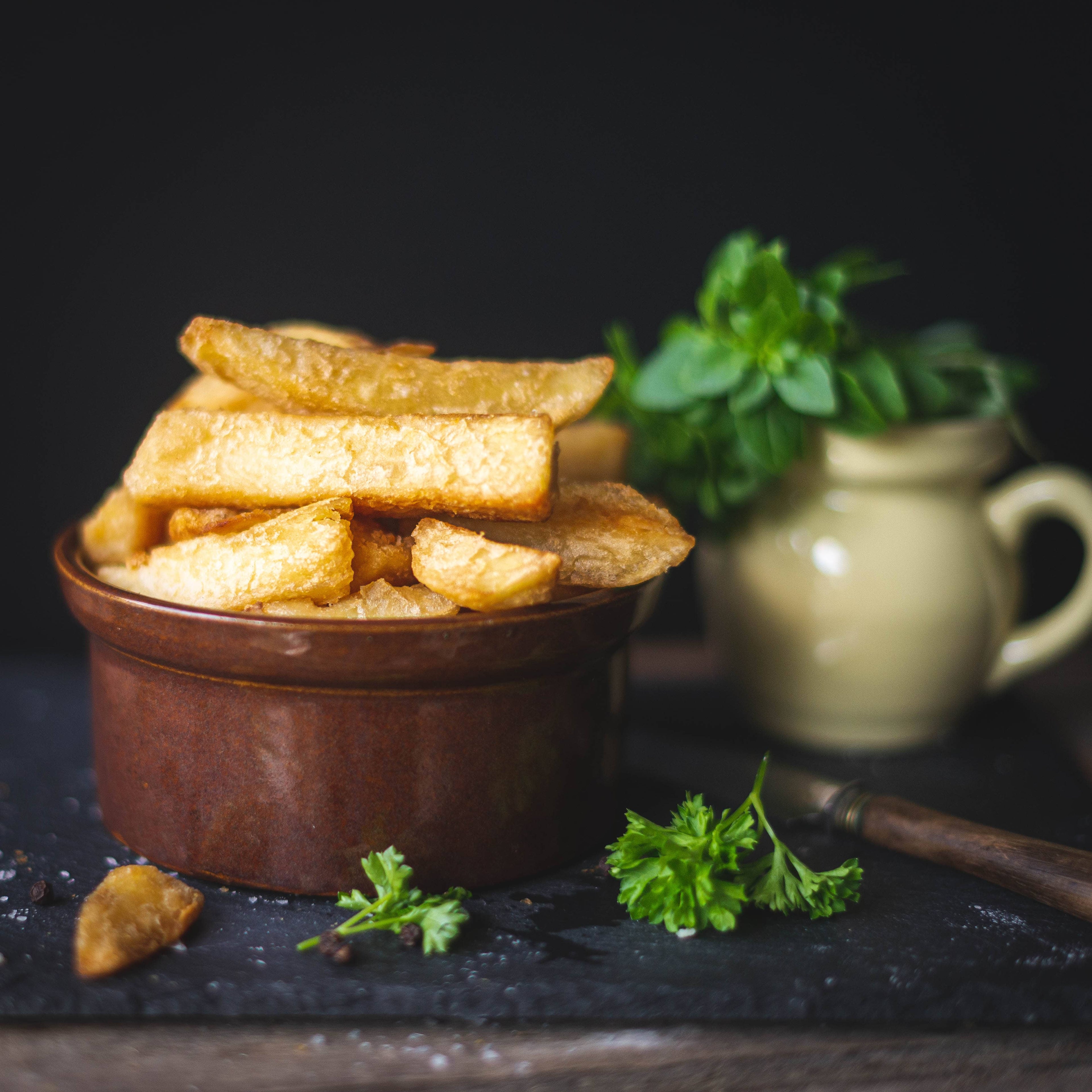 Mud Pies Beer Battered Chips in a brown ceramic bowl rest on a dark surface, with parsley and a cream jug of herbs behind.