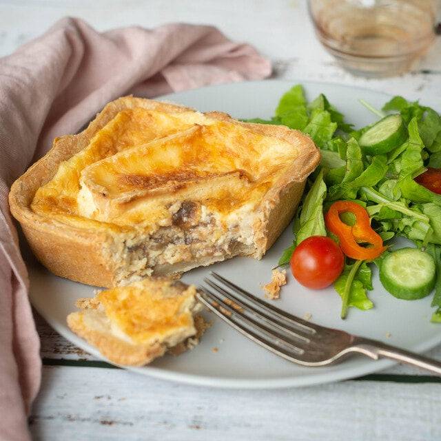 Mud Pies Red Onion Marmalade and Brie Quiche sits on a grey plate with arugula, cucumber, cherry tomatoes, and yellow pepper salad; a fork, napkin, and glass complete the scene.
