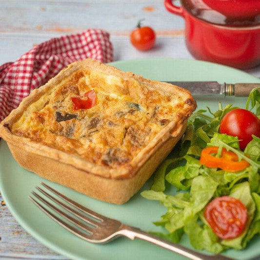 The Mud Pies Roasted Vegetable and Feta Quiche with golden crust sits on a mint-green plate with salad, beside cutlery and red checks.