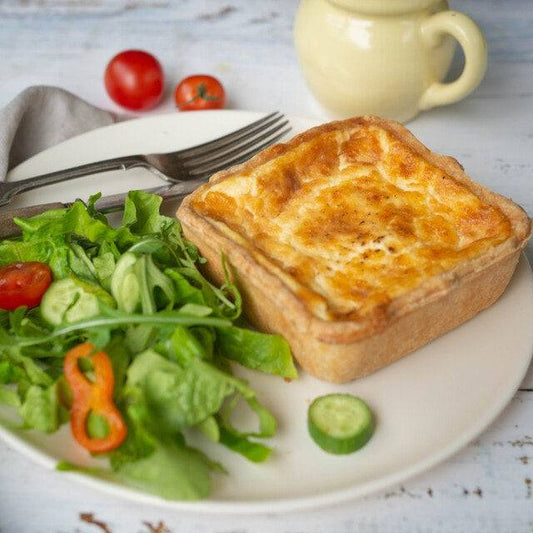 Mud Pies Quiche Lorraine, golden-brown and square, rests on a white plate beside a fresh salad with cutlery and two tomatoes.