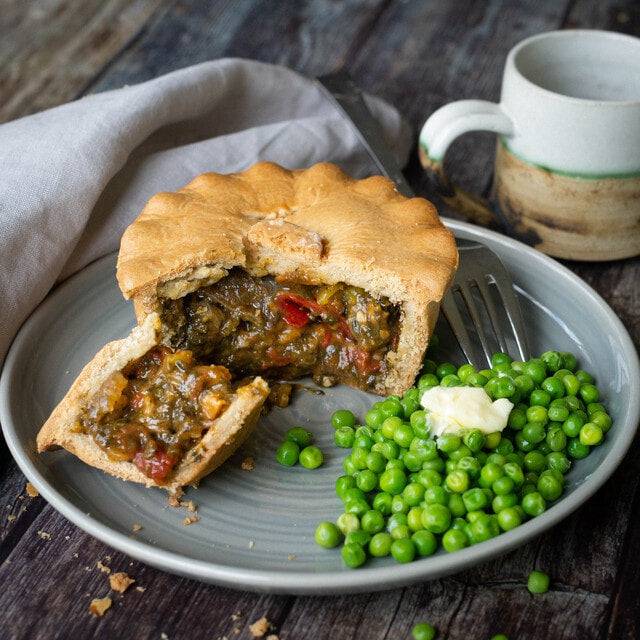 Mud Pies Squash, Spinach and Feta Pie vegan, golden-brown, sits on a grey plate with buttered peas, fork, mug and napkin nearby.