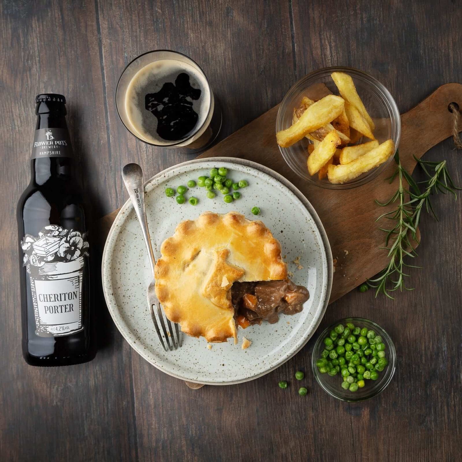 Mud Pies Steak and Ale Pie, sliced open on rustic wood, served with chunky chips, peas, rosemary sprig, fork and dark beer.