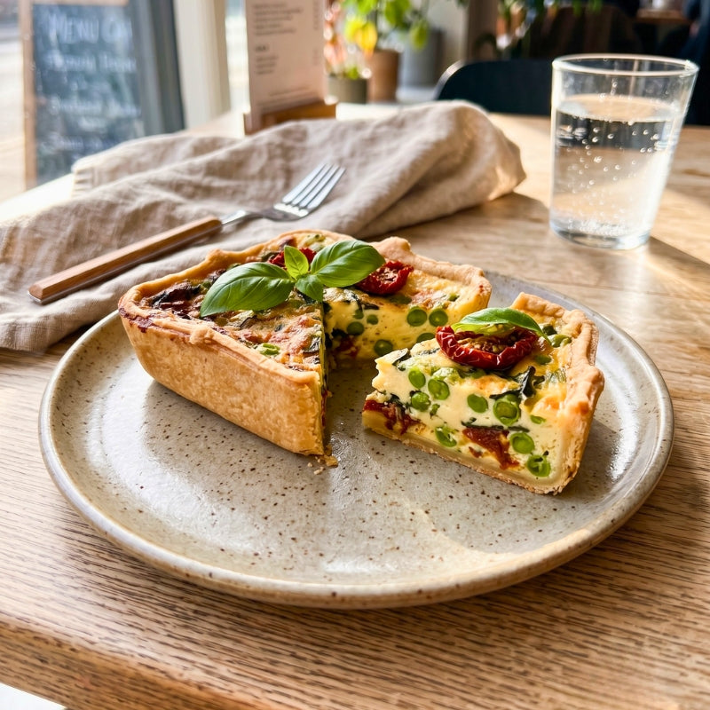 A slice of Mud Pies Pea, Semi-Dried Tomato Twineham Grange Quiche sits in a golden, buttery shortcrust pastry and is topped with fresh basil. The quiche is presented on a beige plate placed on a wooden table. Next to the handmade vegetarian quiche are a fork, napkin, and glass of water. In the softly blurred background, a menu and a plant can be seen.