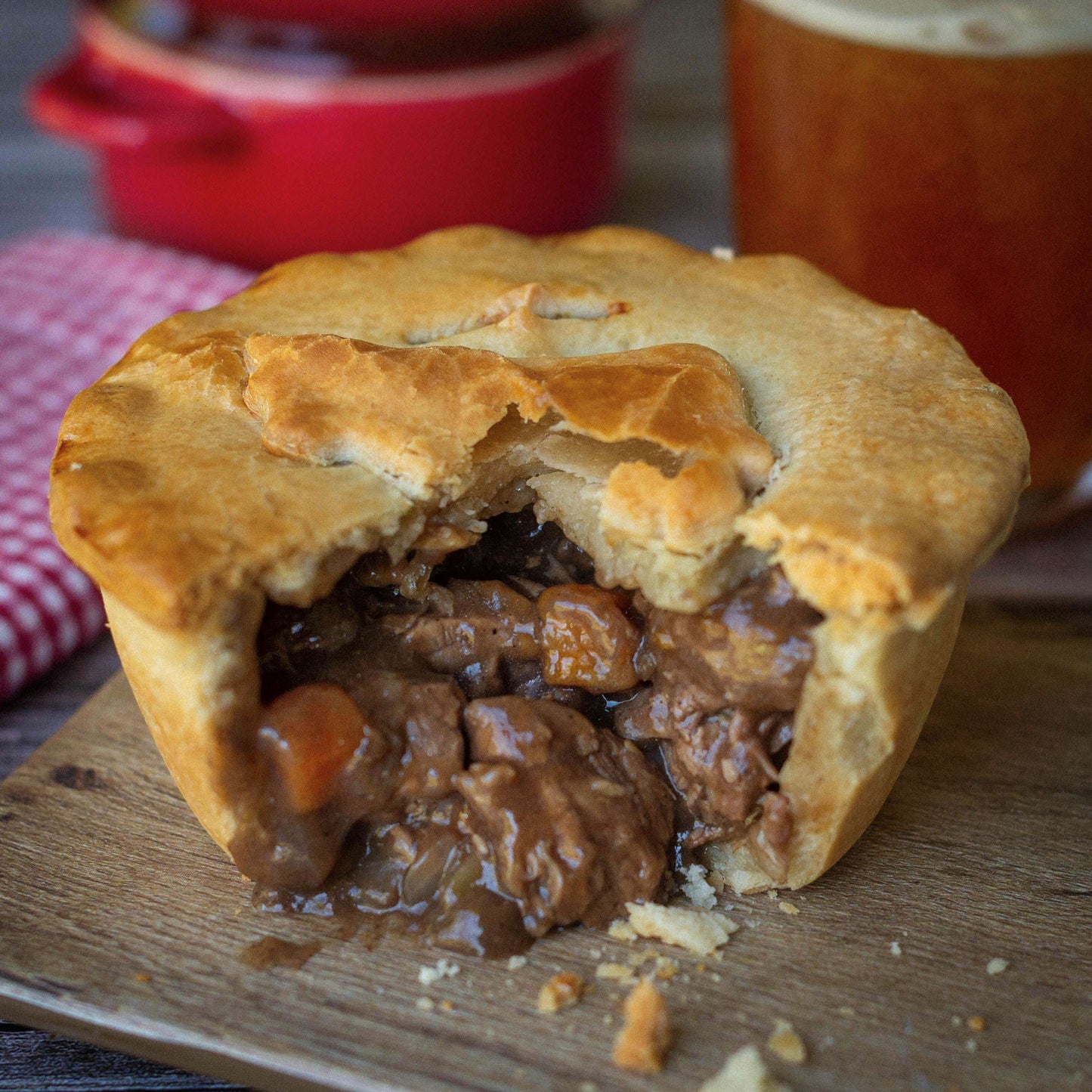 A golden-brown pie from the Mud Pies Ultimate Variety Christmas Gift Box on a wooden board, filling exposed, beer and cloth behind.