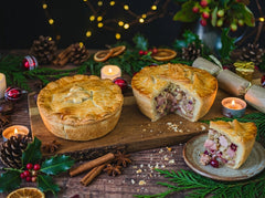A whole and a sliced Mud Pies Christmas In A Pie — Turkey and Ham Christmas Pie with pastry tree decorations sit on a wooden board, with a single slice on a plate; the filling shows chunks of meat, vegetables, and cranberries.