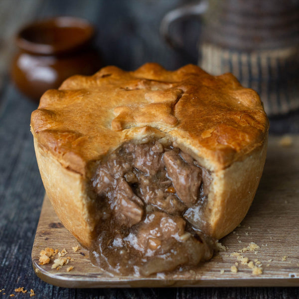 Mud Pies Lamb, Redcurrant and Rosemary Pie, golden-brown with a cut crust, reveals hearty filling on a wooden board; rustic setting.
