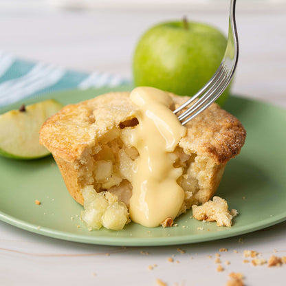 Mud Pies Apple Pie with Cheddar Cheese Crust on a green plate, sliced to show soft filling, topped with custard; apples and crumbs behind.