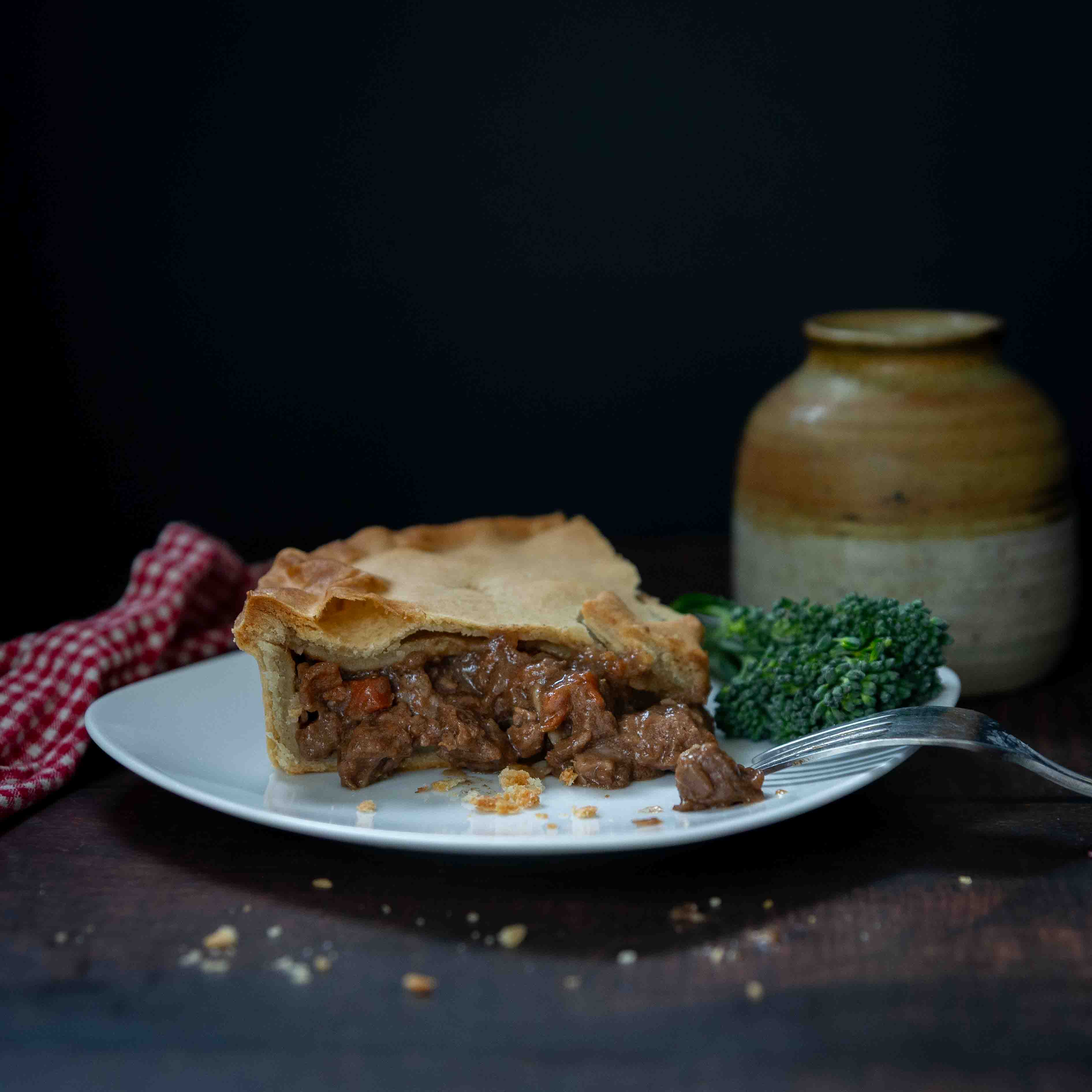 A golden, butter-rich Mud Pies Steak and Ale Family Pie on a white plate with broccoli, a fork, rustic vase and red-checkered cloth.