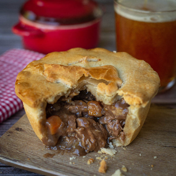 Mud Pies’ Steak and Ale Pie shows its golden crust split to reveal hearty British beef and veg, on a board with beer and red towel.