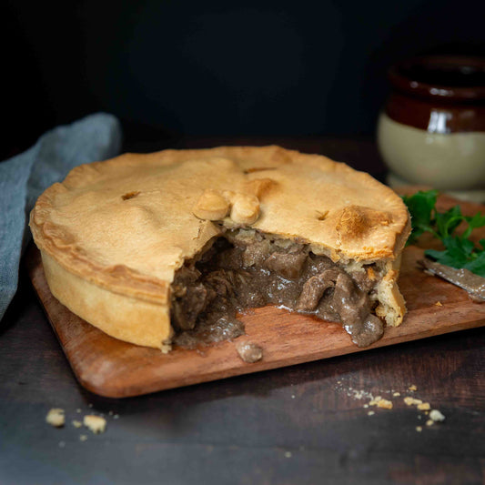 A golden Mud Pies Steak and Ale Family Pie sits on a wooden board, one slice cut to show chunky beef. Crumbs and parsley nearby.