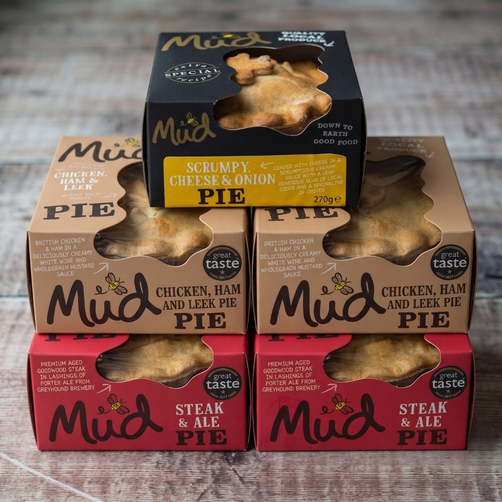 Five Mud Pies brand savoury handmade pies are stacked on a wooden surface: one Scrumpy at the top, two Cheese Onion, one Chicken Ham Leek, and one Steak Ale at the bottom. Each pie comes in vibrant cardboard packaging with a clear window showing the golden crusts. From Britain’s most awarded pie maker, this Pie Taster Box — 5 Best Sellers invites you to try their finest flavours, thoughtfully crafted.
