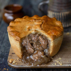 A golden-brown Mud Pies Lamb, Redcurrant Rosemary Pie on a wooden board; flaky crust, tender lamb in gravy; pottery behind.