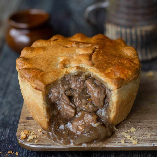 A golden-brown Mud Pies Lamb, Redcurrant Rosemary Pie on a wooden board; flaky crust, tender lamb in gravy; pottery behind.
