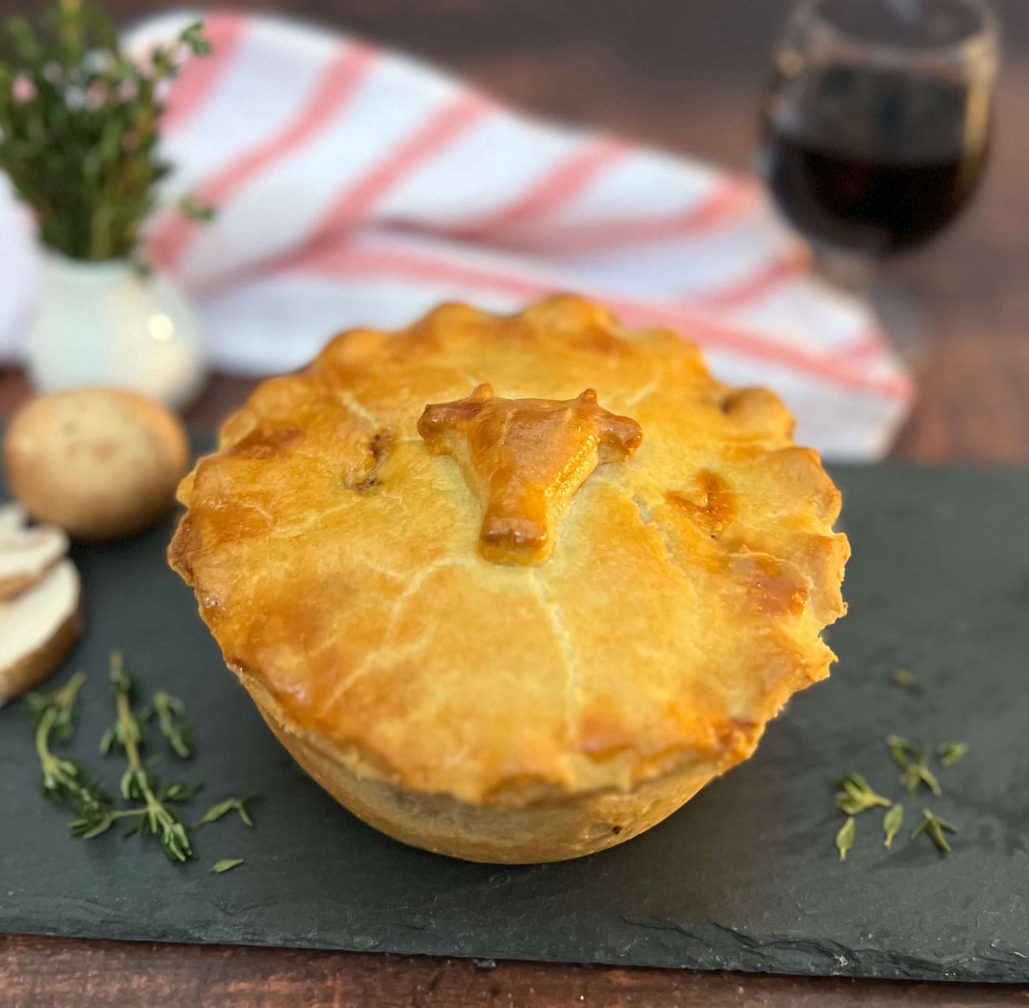 Beef Bourguignon Pie by Mud Pies with a golden shortcrust, bull’s head motif, sits on slate near thyme, mushroom, herbs, wine.