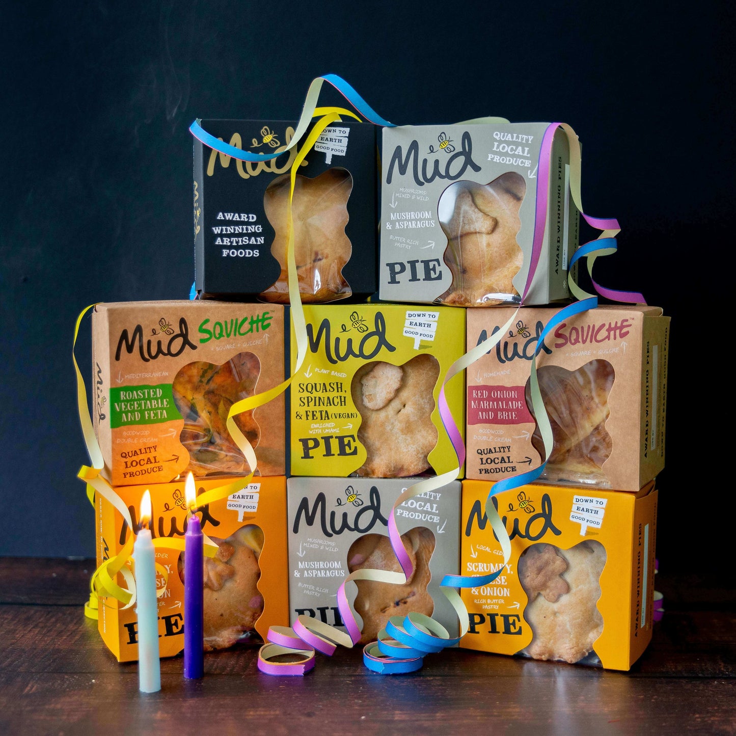 Mud Pies’ Best Ever Veggie Gift Box: savoury pies in vibrant boxes tied with yellow, blue, and pink ribbons, with white and purple candles in front. Squash and mushroom flavours are featured.