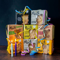 Mud Pies’ Best Ever Veggie Gift Box: savoury pies in vibrant boxes tied with yellow, blue, and pink ribbons, with white and purple candles in front. Squash and mushroom flavours are featured.
