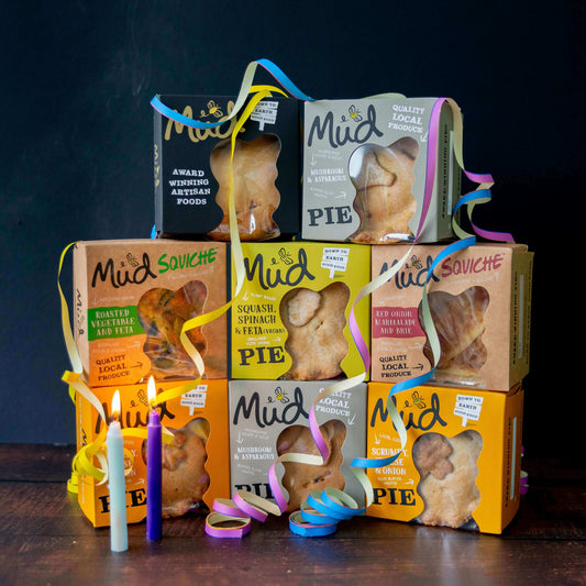 Mud Pies’ Best Ever Veggie Gift Box: savoury pies in vibrant boxes tied with yellow, blue, and pink ribbons, with white and purple candles in front. Squash and mushroom flavours are featured.