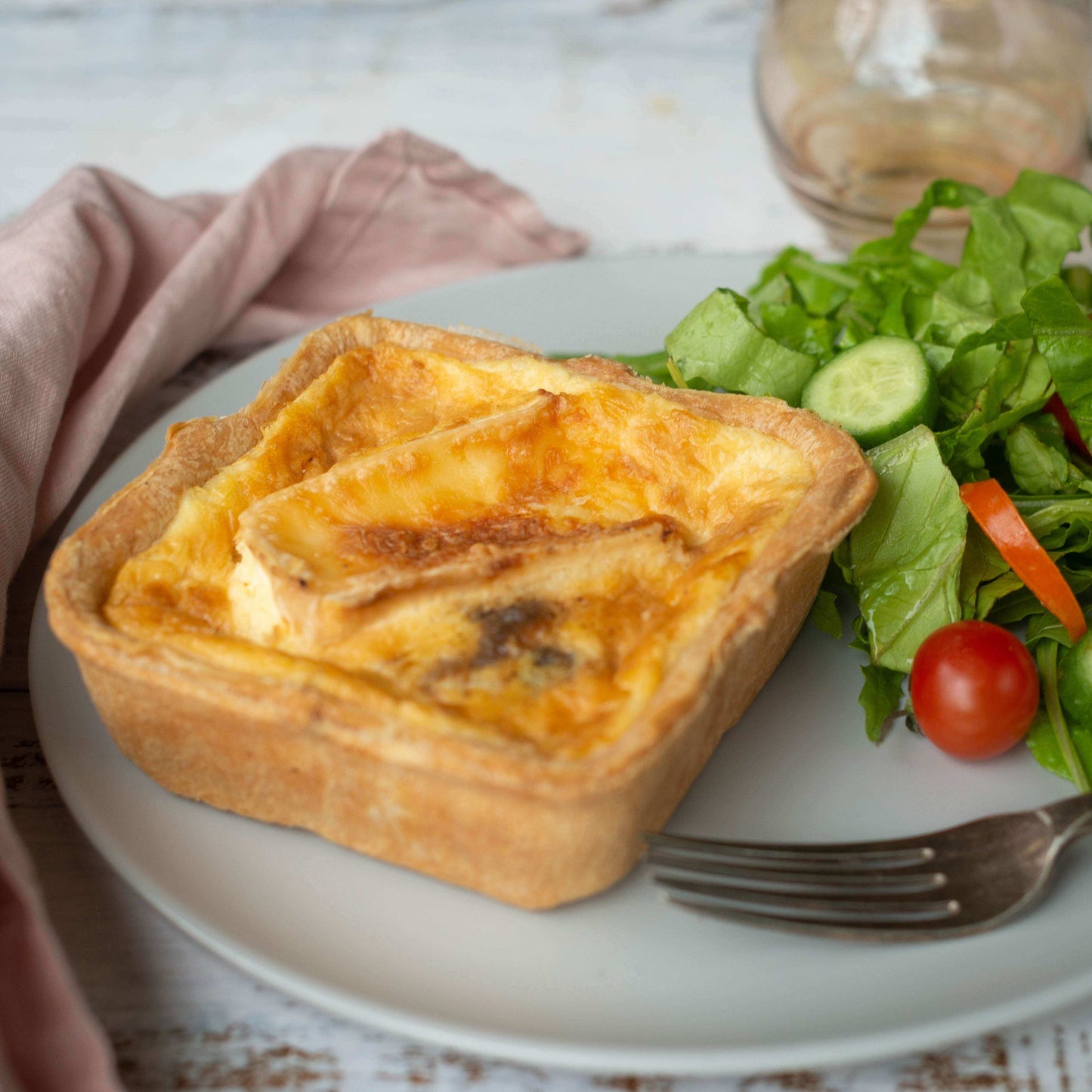 Mud Pies' Best Ever Veggie Gift Box slice with a golden flaky crust and melted cheese on a white plate, beside salad, fork and pink napkin on rustic wood.
