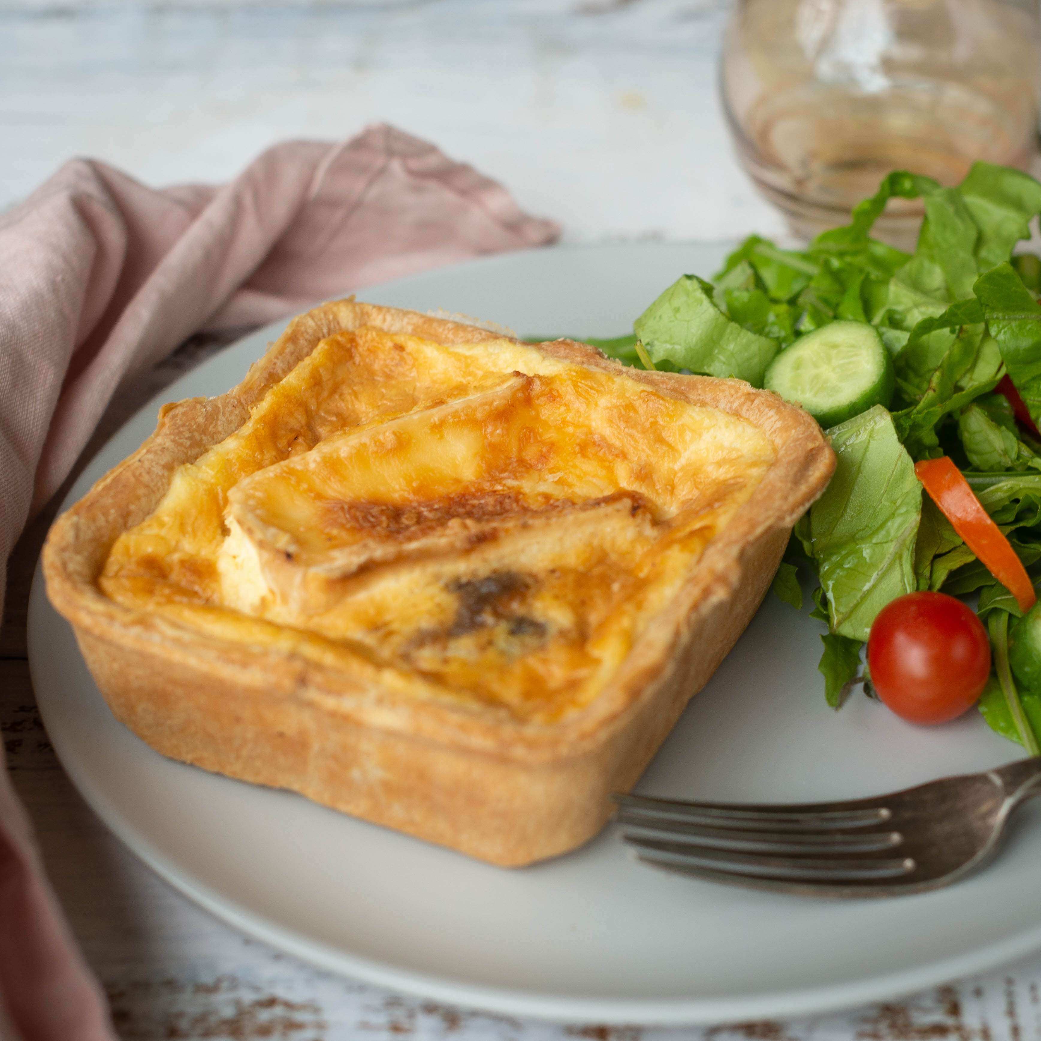 Mud Pies' Best Ever Veggie Gift Box slice with a golden flaky crust and melted cheese on a white plate, beside salad, fork and pink napkin on rustic wood.