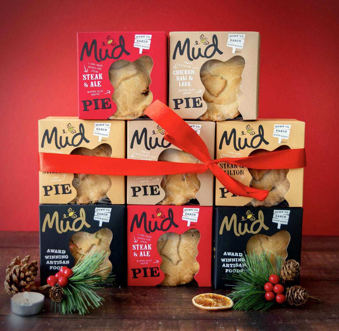 A Best Sellers Christmas Pie Gift Box by Mud Pies features a stack of nine handmade pies tied with a red ribbon on a dark wooden surface, with windowed boxes revealing golden pastries, flavour labels, pinecones, and holly.
