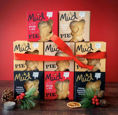 A Best Sellers Christmas Pie Gift Box by Mud Pies features a stack of nine handmade pies tied with a red ribbon on a dark wooden surface, with windowed boxes revealing golden pastries, flavour labels, pinecones, and holly.