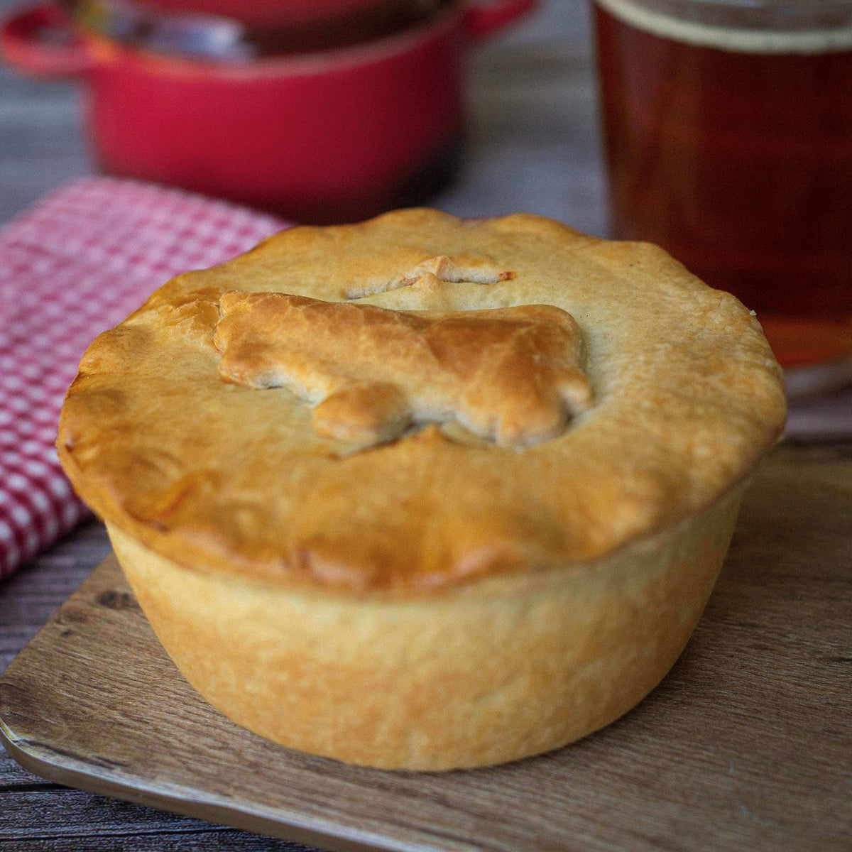 A golden-brown Best Sellers Christmas Pie Gift Box by Mud Pies featuring a decorative pastry cow on top, served on a wooden board with a red checkered napkin, a small red pot, and a glass of amber liquid in the background.