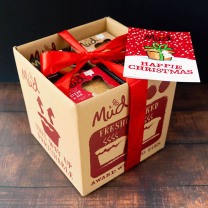 A Best Sellers Christmas Pie Gift Box from Mud Pies filled with handmade meat pies, tied with a large red ribbon, featuring a festive red greeting card on top that reads HAPPIE CHRISTMAS.