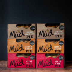 Six Mud Pies Best Sellers Pie Box 6 Pack cartons sit in two neat columns on wood, each with a bold “great taste” label, against black.