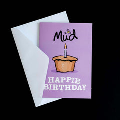 Birthday Greetings Card by Mud Pies, partially beneath a white envelope, shows a lit cupcake, "Mud" with a bee, and bold "HAPPIE BIRTHDAY".