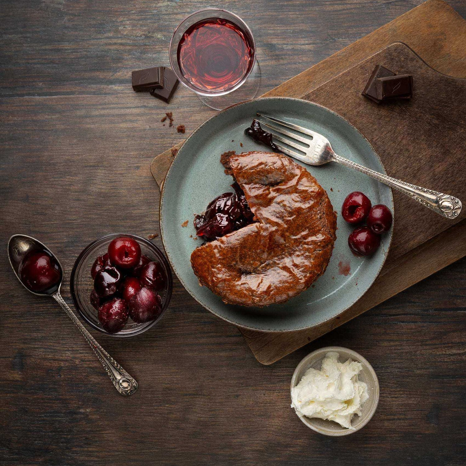 Black Forest Gateau Pie by Mud Pies, with a slice out, rests on a plate with cherries and a fork, ringed by chocolate, cream and wine.