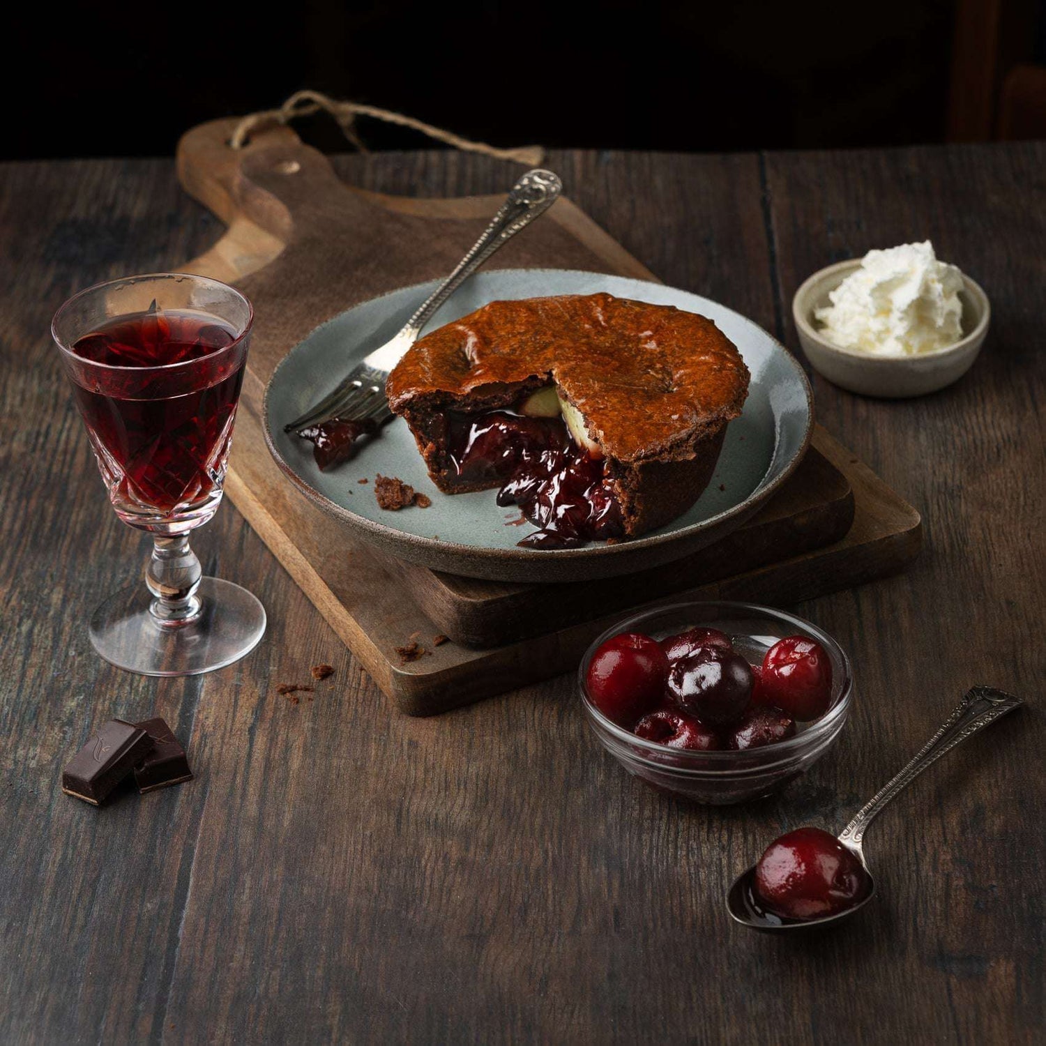 Mud Pies’ Black Forest Gateau Pie sits on a plate atop a rustic wooden table, with its cherry filling peeking from the crust. Nearby are a glass of red wine, whipped cream, chocolate pieces, fresh cherries and a spoon.