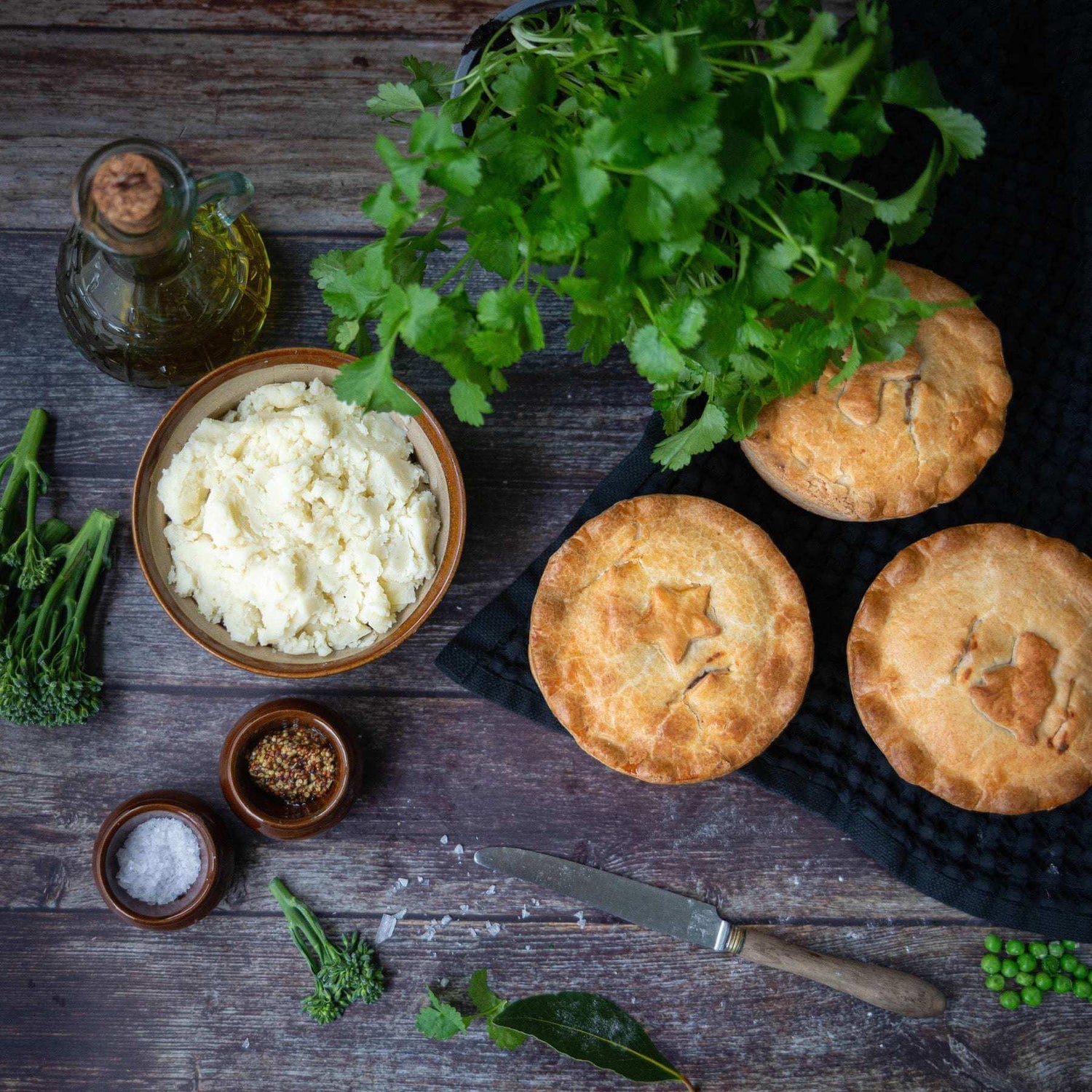 Golden Mud Pies sit on a black cloth with Buttery Mashed Potato. Fresh coriander, olive oil, salt, mustard, broccoli, peas and a knife are spread across a rustic wooden table.