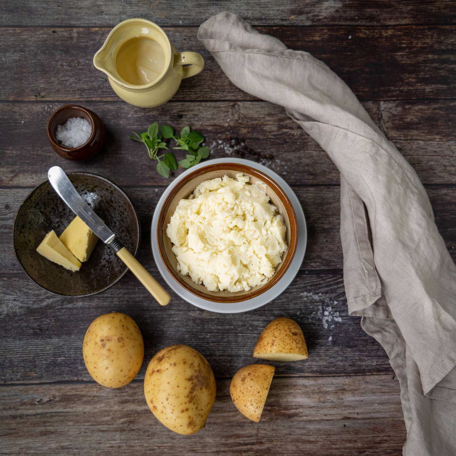 Mud Pies Buttery Mashed Potato in a bowl on a rustic wooden table, with whole and cut potatoes, butter, fresh herbs, and a beige cloth.