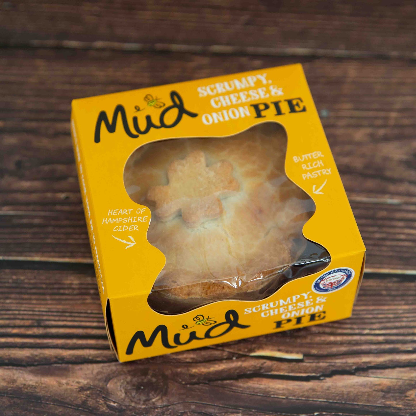 Mud Pies Scrumpy Cheese and Onion Pie (Cheese, Onion and Potato Pie) in a yellow box with a clear window, highlighting buttery pastry and Hampshire cider, on wood table.