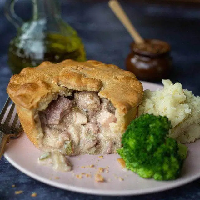 Mud Pies’ Chicken, Ham and Leek Pie in golden shortcrust pastry, with mash and broccoli on a pink plate; oil bottle, jar behind.