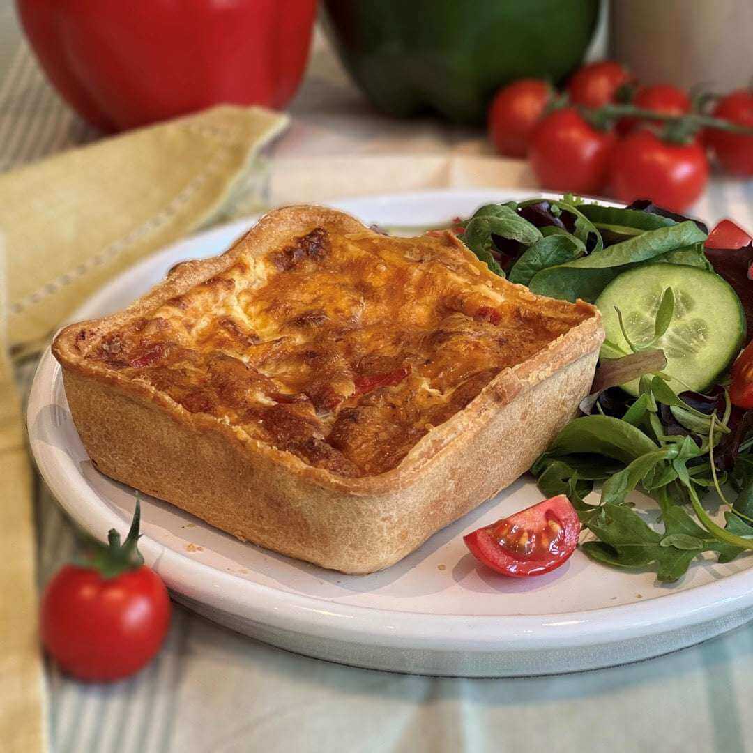 Mud Pies Roasted Pepper and Chorizo Melt Quiche, square-cut, on a white plate with mixed leaf salad, cucumber and cherry tomatoes.