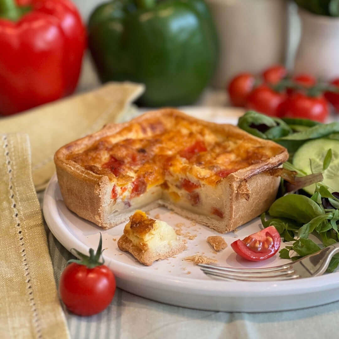 Mud Pies Roasted Pepper and Chorizo Melt Quiche, square with golden crust and red pepper pieces, sits on a white plate with a slice removed, showing creamy filling; fresh greens and tomatoes on the side.