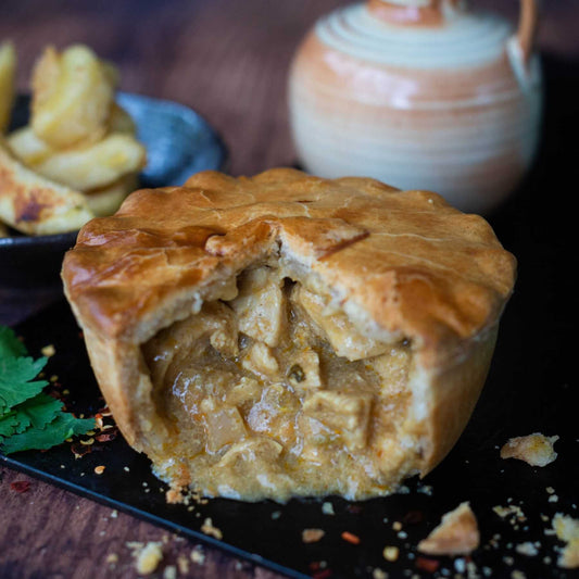 Mud Pies Chip Shop Chicken Curry Pie, golden and flaky, sits sliced open to reveal rich filling; potato wedges and a jug behind.