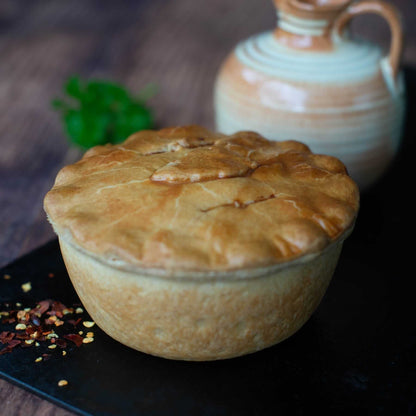 Mud Pies Chip Shop Chicken Curry Pie, golden and flaky, rests on a black surface with red pepper flakes, jug and fresh parsley behind.