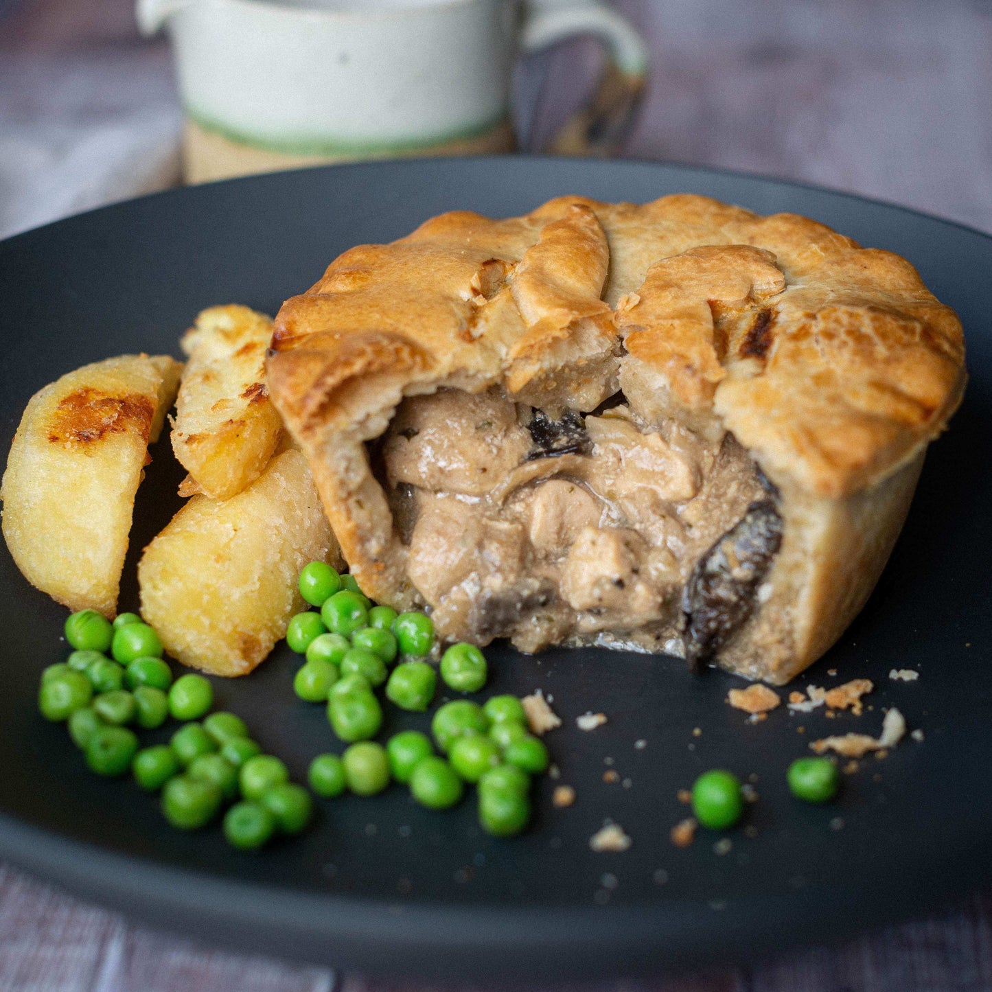 Mud Pies' "The Complete Gift Box" pie, golden and flaky, reveals creamy meat filling; served with roast potatoes, peas, and a blurred mug.