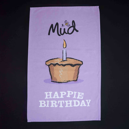 The Mud Pies Tea Towel Birthday is a purple cotton towel with a candle-topped cupcake, bee, “Mud”, and “HAPPIE BIRTHDAY” text.
