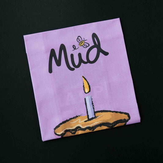 Mud Lavender Tea Towel: folded, with "Mud" in black script, a bee over the 'u', and a pie topped by a lit candle, set against black.