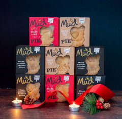 A Luxury Christmas Collection Gift Box by Mud Pies, with steak and ale pies in two rows, adorned with candles, pinecone, berries, and a red ribbon on a dark backdrop.