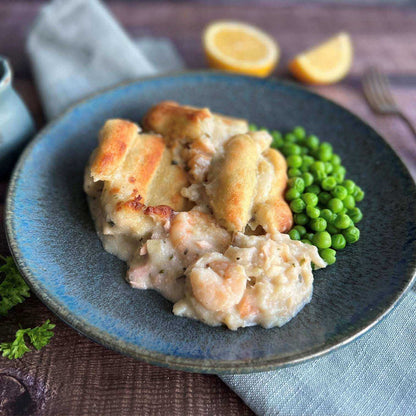 Mud Pies Luxury Fish Pie sits on a blue plate, golden-topped with seafood showing, served beside vibrant peas and lemon halves.