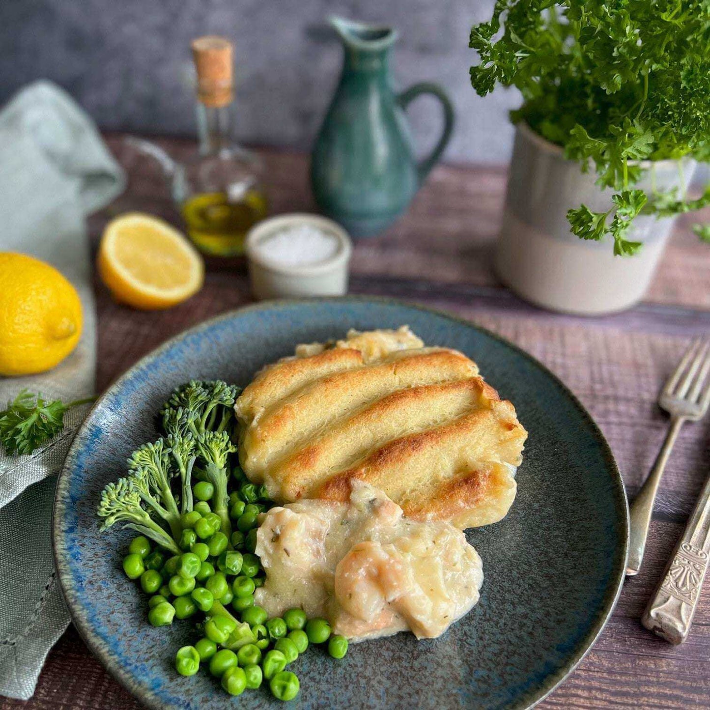 Mud Pies’ Luxury Fish Pie on a blue plate, creamy with smoked fish and mash, with broccoli and peas; lemon and herbs behind.