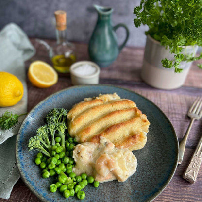 Mud Pies’ Luxury Fish Pie on a blue plate, creamy with smoked fish and mash, with broccoli and peas; lemon and herbs behind.