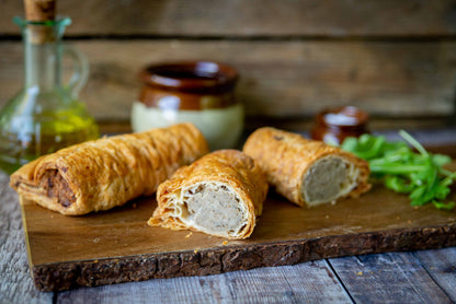 Mud Pies Luxury Sausage Rolls—three golden-brown pastries, one cut open to show the pork filling—on a rustic wooden board, herbs and jars behind.