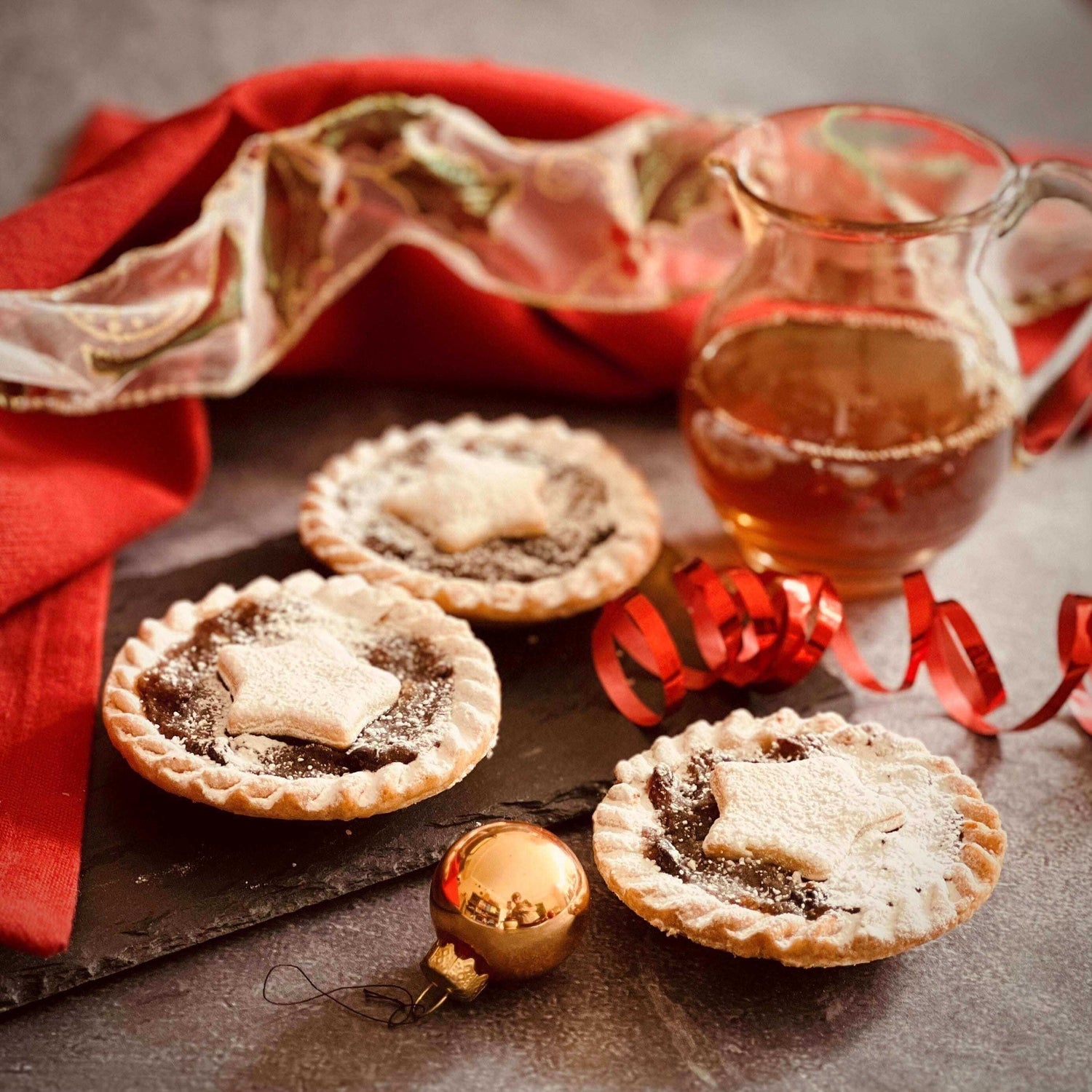 Three Mud Pies Brandy Laced Luxury Mince Pies with pastry stars sit on slate, beside a jug of brandy and festive red-gold decor.